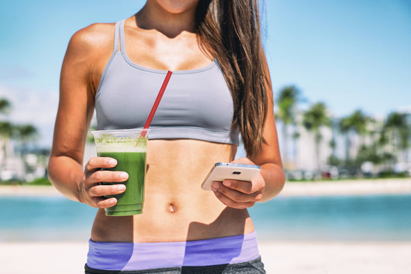 Young woman with a flat stomach Googling "What is a gut cleanse?" and holding a green smoothie on a beach